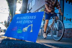 An image of a cyclist on the sidewalk, next to a sign that says "Businesses Are Open"
