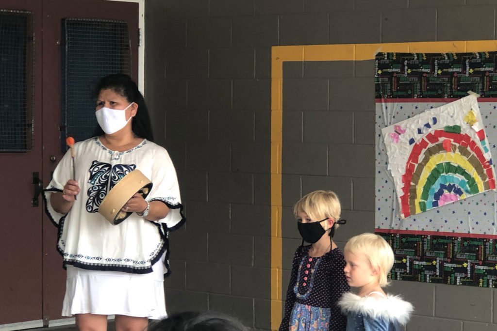 An outdoor image of a childcare worker playing a traditional hand drum and standing together with two young children. One child and the childcare worker are wearing masks.