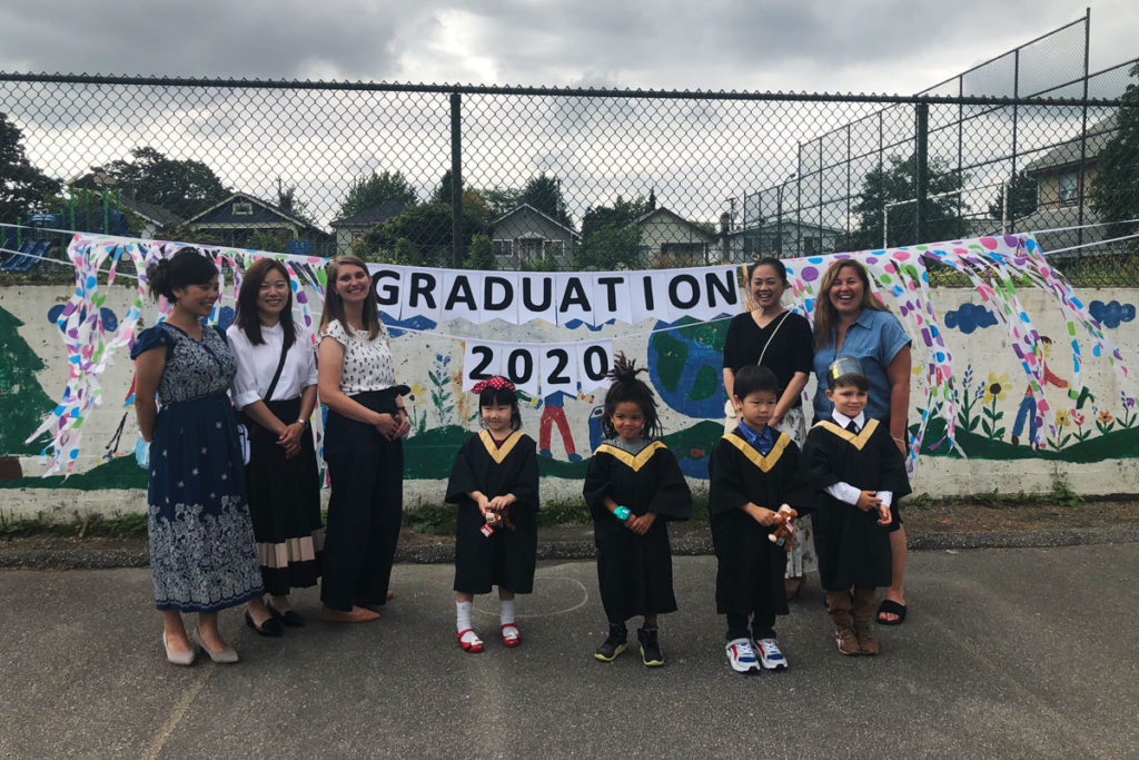 An outdoor image of a group of children wearing graduation gowns and caps, along with childcare workers standing in front of a banner that says "Graduation 2020"